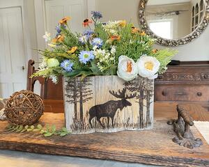 A wood moose box made with upcycled gray wood. A moose standing in the woods is wood-burned onto the front. It sits on a dining room table. 
